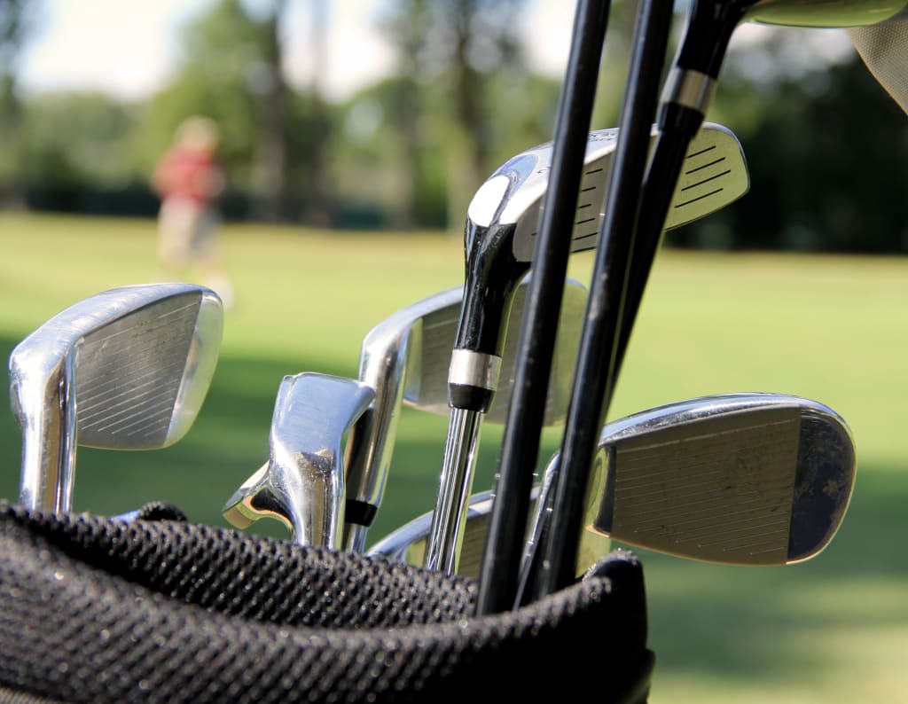 Close-up of golf clubs, including some of the most forgiving golf irons, in a bag on a golf course with a blurred figure in the background.